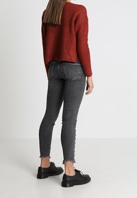 Roter Strickpullover mit herabgesetzten Schultern, kombiniert mit hochgeschnittenen schwarzen Jeans mit fransigen Säumen und Knopfdetails, getragen mit schwarzen Schuhen.