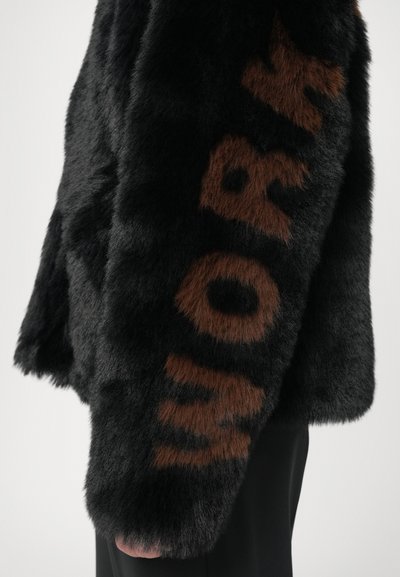 Sort faux fur jakke med en brun "WORK" tekstdetalje, blød tekstur og rund silhuet.