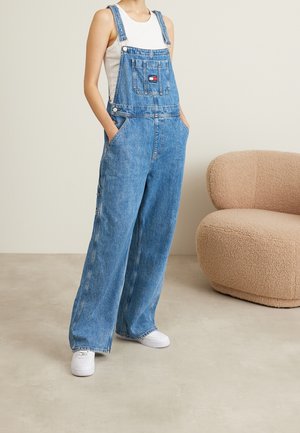 Person iført blå denim-overalls med vidde ben og hvide sneakers, stående nær en beige tekstureret lænestol på et lyst gulv.