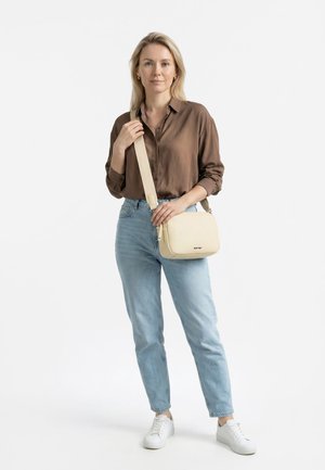 Kvinde med langt blondt hår iført en brun skjorte med knapper, lyseblå jeans, hvide sneakers og en cremefarvet skuldertaske.