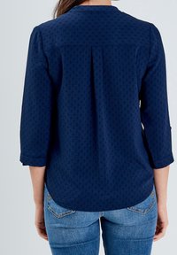 Femme portant un chemisier bleu marine à manches longues avec une texture à pois subtils et un jean bleu, vue de dos sur un fond uni.