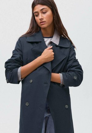 Donna che sistema un cappotto doppiopetto blu scuro con le maniche arrotolate, indossato sopra un maglione grigio e jeans azzurri chiari.