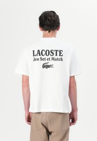 Balts kokvilnas T-krekls ar melnu tekstu "LACOSTE Jeu Set et Match" un grafisku krokodila logo aizmugurē. Īsas piedurknes, brīvs piegrieziens.