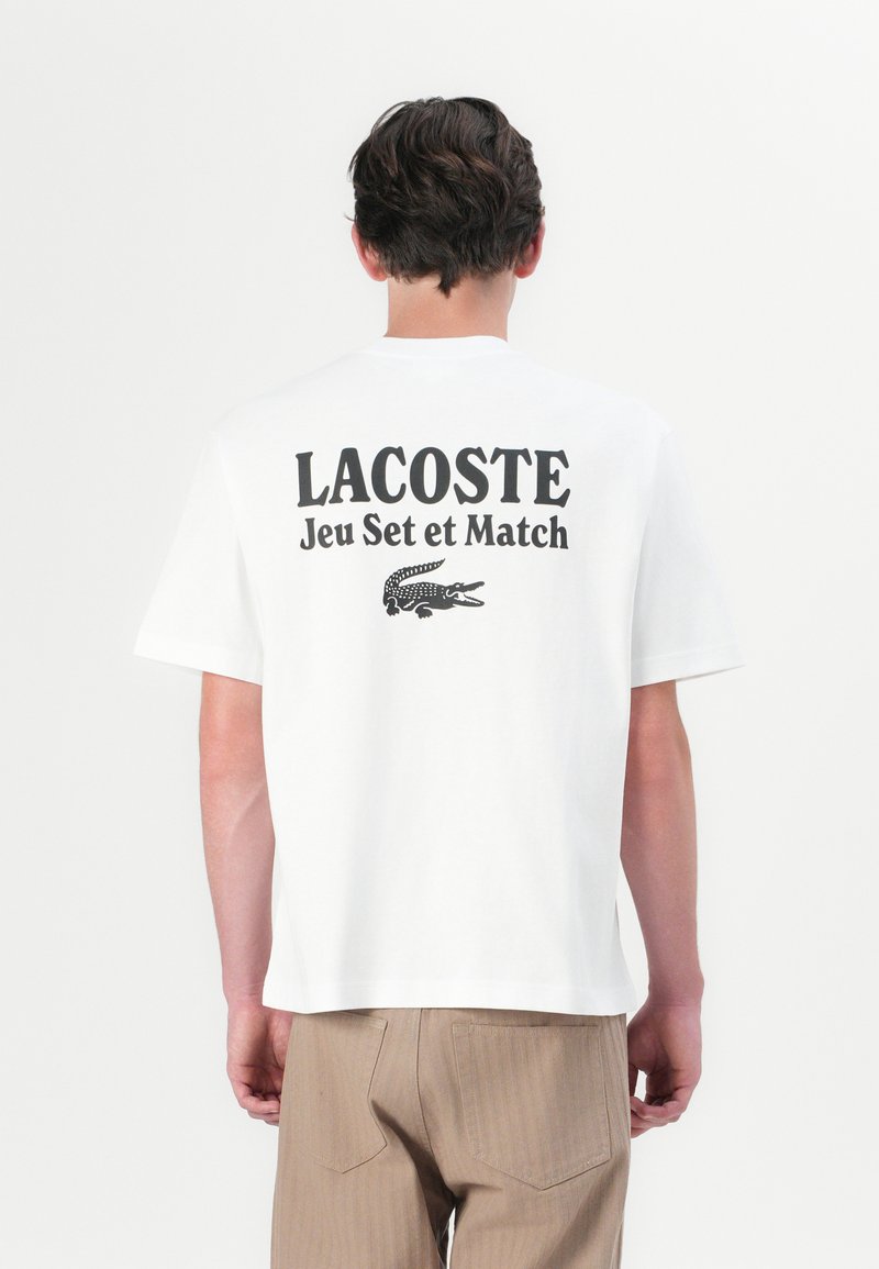 Balts kokvilnas T-krekls ar melnu tekstu "LACOSTE Jeu Set et Match" un grafisku krokodila logo aizmugurē. Īsas piedurknes, brīvs piegrieziens.