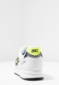 Scarpa atletica bianca con accenti grigi e giallo neon, tomaia in rete, colletto imbottito e suola in gomma. Presenta il logo "asics" sul tallone.