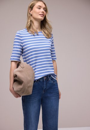 Frau mit langen blonden Haaren, die ein blau-weiß gestreiftes Hemd und blaue Jeans trägt und in einer Hand eine beige Wildlederjacke hält.