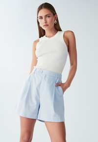 Světle modré šortky s vysokým pasem, sklady a bočními kapsami, spárované s bílým žebrovaným tílkem. Hladká tkanina a knoflíkový detail na pase.