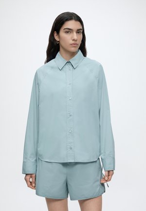 Woman with long dark hair wears light blue button-up shirt and matching shorts against plain white background.