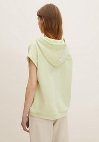 Top sin mangas capucha de color verde claro con un corte relajado, textura suave y mangas enrolladas. Diseño simple sin patrones ni adornos visibles.