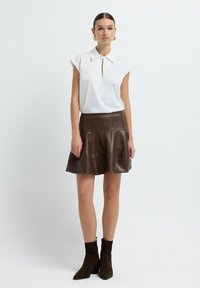 Blouse blanche à manches courtes avec un col et une ouverture devant. Jupe évasée en faux cuir marron avec des coutures. Bottines en suède marron à chevilles.