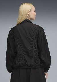 Black lightweight jacket with a gathered waistband, textured fabric, and puffed sleeves. Features a classic collar and seam detailing at the back.