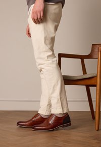Braune Lederdressschuhe mit strukturiertem Finish, ausgestattet mit Schnürdesign, schwarzen Mesh-Akzenten und einer kontrastierenden blauen Sohle.