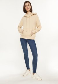Beige kapucnis pulóver elülső zsebbel, állítható húzózsinórral és bordázott mandzsettákkal, kék skinny farmernadrággal és fehér tornacipővel párosítva.