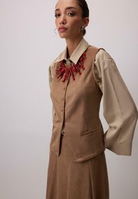 Nő bézs blúzban, barna suede mellényben és szoknyában, vastag, vörös korall stílusú nyaklánccal kiegészítve, sima háttér előtt áll.