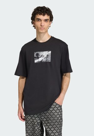 Svart t-shirt med kort ärm som har ett grafiskt tryck av en sneaker på en vit rektangulär bakgrund, bärs med mönstrade grå byxor.