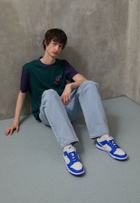 Blue and white low-top sneakers with suede and leather textures, paired with light blue jeans and a green-purple color-blocked shirt.