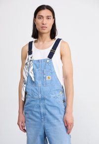 Vaaleansiniset farkkuhaalarit, joissa on tummansiniset hihnat, edessä tasku ja etiketti. Valkoinen bandana on sidottu sivulle.