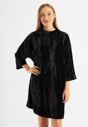 Schwarzes Samtkleid mit lockerer Passform, 3/4-Ärmeln und hohem Kragen. Der Stoff hat eine gerippte Textur und einen subtilen Glanz.