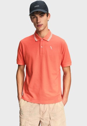 Giovane uomo con polo color corallo bordata di bianco, pantaloncini cargo beige e cappellino da baseball nero Aigle, in piedi con le mani nelle tasche.