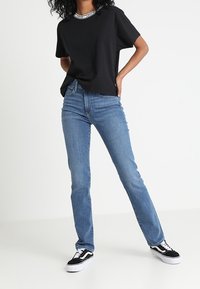 Osoba oblečená v černém tričku, světle modrých slim-fit džínech a černo-bílých teniskách stojící s jednou rukou na boku.
