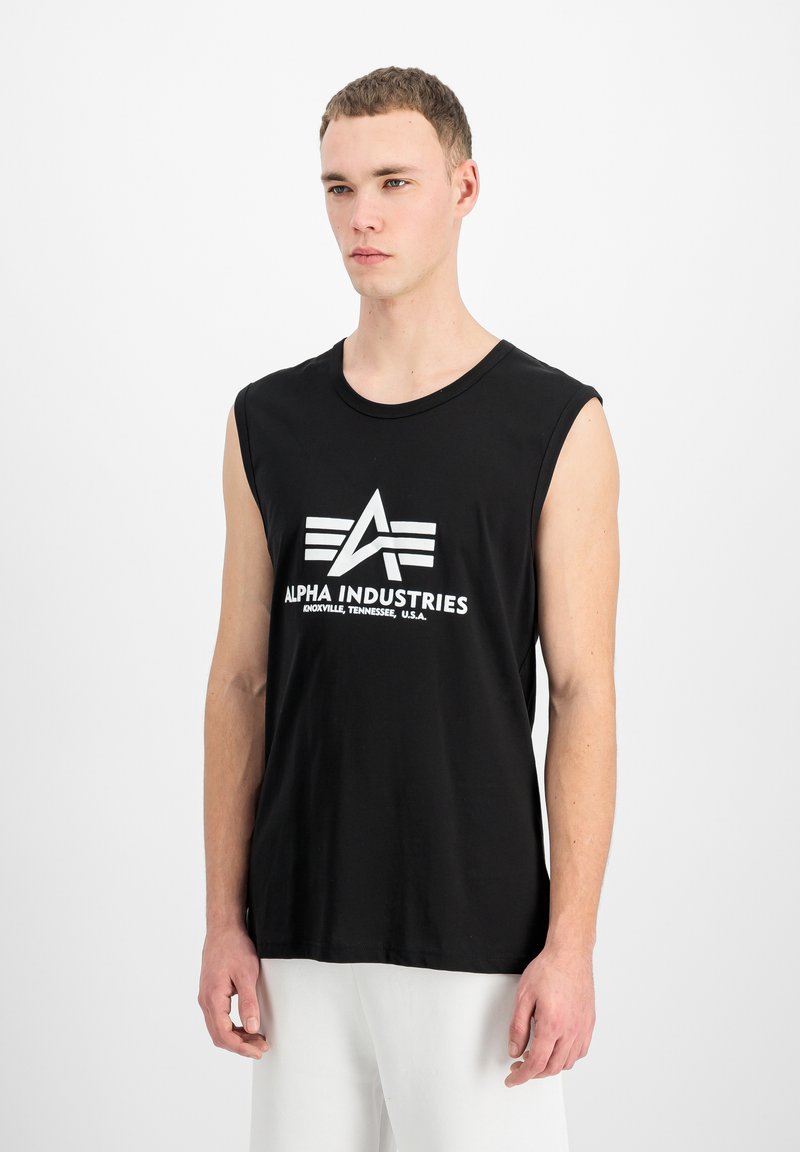 Jeune homme aux cheveux courts portant un débardeur noir avec le logo "Alpha Industries Knoxville, Tennessee, U.S.A." et un pantalon blanc.