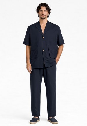 Mężczyzna w granatowej marynarce z krótkim rękawem z dwoma przednimi kieszeniami, dopasowanych spodniach i granatowych butach wsuwanych, stojący na białym tle.