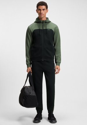 Black and green zip-up hoodie with a hood, paired with matching black joggers. Dark bag with shoulder strap held in hand.