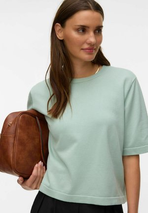 Donna con lunghi capelli castani indossa un maglione verde chiaro a maniche corte, tiene uno zaino in pelle marrone sotto il braccio e guarda di lato.