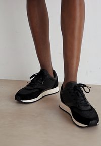 Svarte sneakers med en blanding av teksturert semsket skinn og glatt lær, med en kontrasterende hvit såle og svarte snørebånd. Accenter inkluderer subtile merkeelementer.