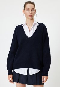 Laivastonsininen neulepusero, jossa on V-pääntie, ribbikuvio ja valuvaa olkasaumat, kerrostettuna valkoisen kauluspuseron päälle ja yhdistettynä laskostettuun hameeseen.