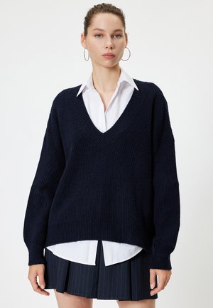 Navy gebreide trui met een V-hals, geribbelde textuur en verlaagde schouders, gelaagd over een witte coll shirt en gecombineerd met een plissérok.