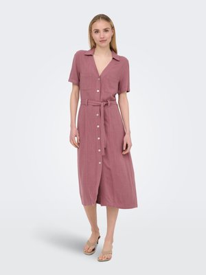 Femme debout portant une robe midi boutonnée de couleur vieux rose, manches courtes, ceinture et sandales à talons beiges sur un fond blanc uni.