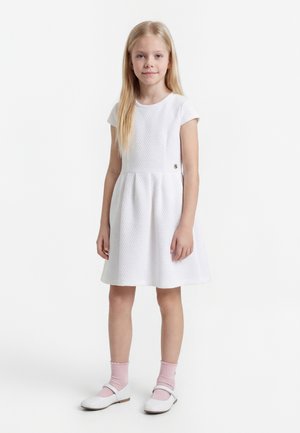 Young girl standing, wearing a white textured dress, pink socks, and white Mary Jane shoes on a plain white background.
