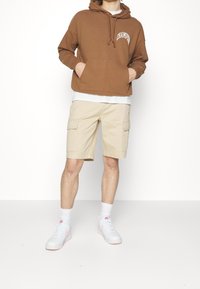 Sudadera con capucha marrón con bolsillo delantero y logo de "Champion", combinada con pantalones cortos cargo beige claro con bolsillos laterales y zapatillas blancas.