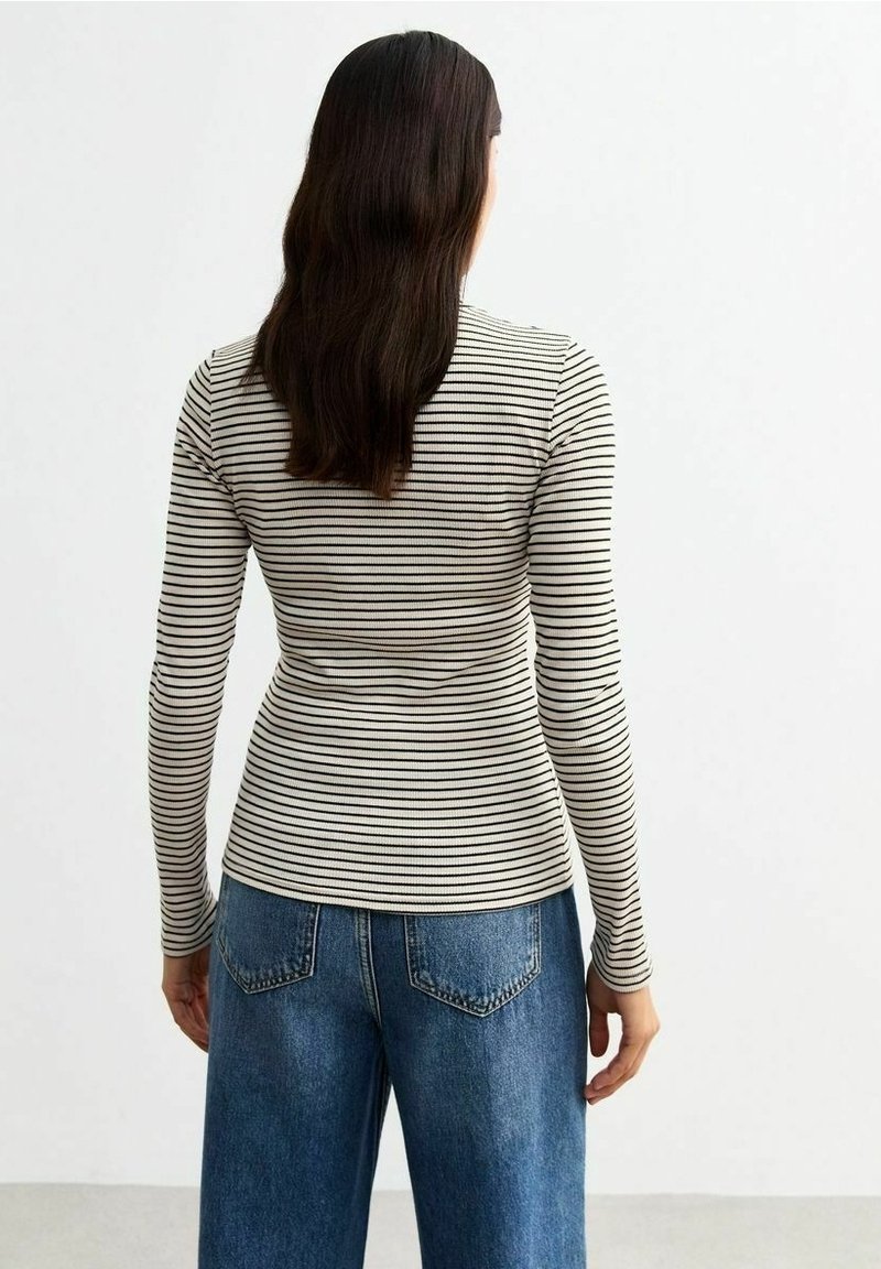 Femme aux longs cheveux foncés portant une chemise à manches longues beige et noire à rayures et un jean bleu, debout de dos sur un fond blanc.