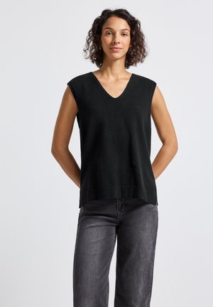 Frau mit lockigem Haar, die einen ärmellosen schwarzen V-Ausschnitt-Pullover und ausgebleichte schwarze Jeans trägt und vor einem hellen Hintergrund steht.
