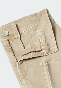 Pantalon en jean beige avec fermeture éclair partiellement ouverte, bouton métal visible, détails de couture et passants pour ceinture sur un fond clair.
