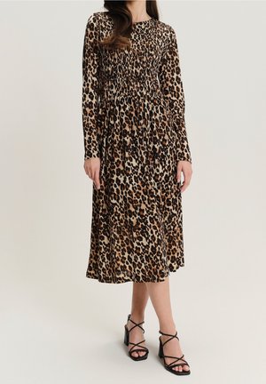 Frau trägt ein langärmliges, knielanges Kleid mit Leopardenmuster, geschmückter Taille und schwarzen Riemchen-Sandalen mit Blockabsatz.