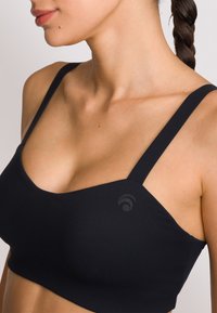 Zwarte sportbeha met brede banden, een gladde textuur en een kenmerkend logo aan de voorkant. Ontworpen voor comfort en steun.