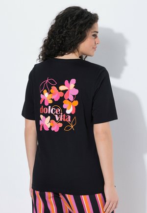 Vrouw met krullend haar die een zwart T-shirt draagt met een roze en oranje bloemenpatroon en de tekst "dolce vita" op de achterkant.