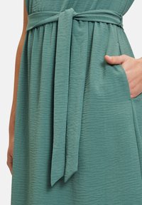 Hellblaues Kleid aus strukturiertem Stoff, mit tailliertem Schnitt und gebundenem Gürtel sowie seitlichen Taschen.