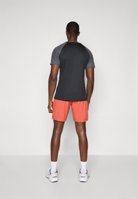 Schwarzes und graues Sportshirt mit kurzen Ärmeln, kombiniert mit korallenfarbenen Shorts. Weiße Sneaker vervollständigen den Look. Einfarbiger Hintergrund.