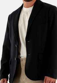 Blazer in misto lana blu navy con design elegante, revers a punta, due bottoni e tasca frontale. Abbinato a una camicia bianca e pantaloni beige.