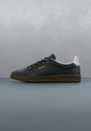 Schwarzer flacher Sneaker mit brauner Sohle, weißem Fersenlaschen, schwarzen Schnürsenkeln und goldenem „BOSS“-Logo an der Seite, vor grauem Hintergrund.