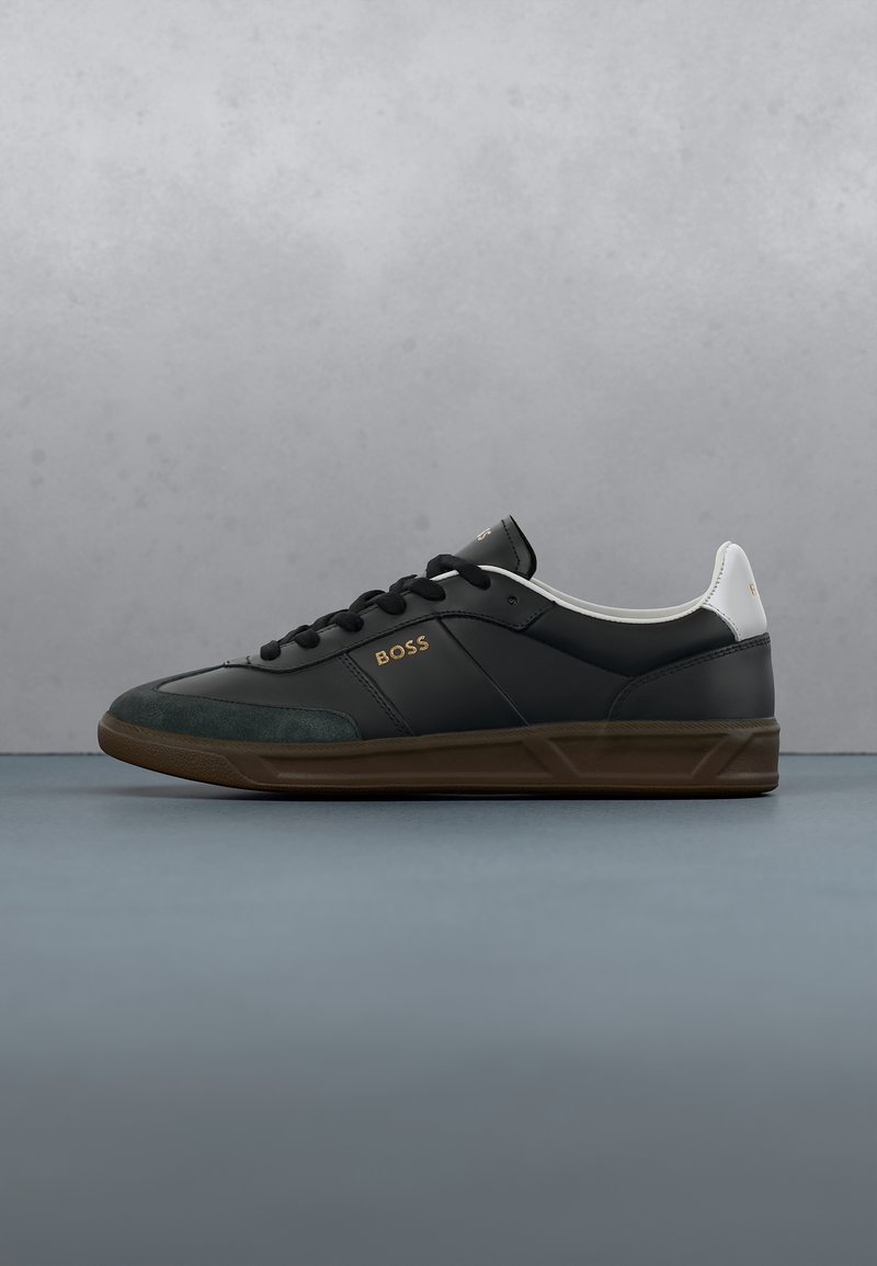 Basket basse noire avec semelle marron, languette de talon blanche, lacets noirs et logo doré « BOSS » sur le côté, sur fond gris.