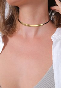 Guldtonet metal choker med en glat, buet stang på en sort snor. Designet er minimalistisk og moderne, hvilede mod bar hud.