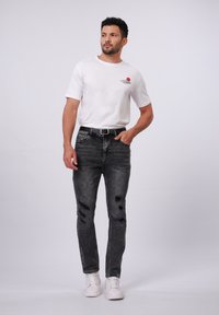 Camiseta blanca de manga corta con un gráfico, pantalones vaqueros negros desgastados con rotos y zapatillas blancas. Conjunto casual con un diseño texturizado.