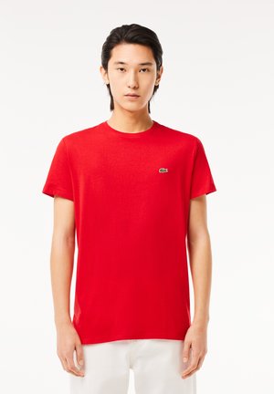 Camiseta de algodón roja con cuello redondo, mangas cortas y un pequeño logo verde de Lacoste en el pecho izquierdo. Textura suave, corte ajustado.
