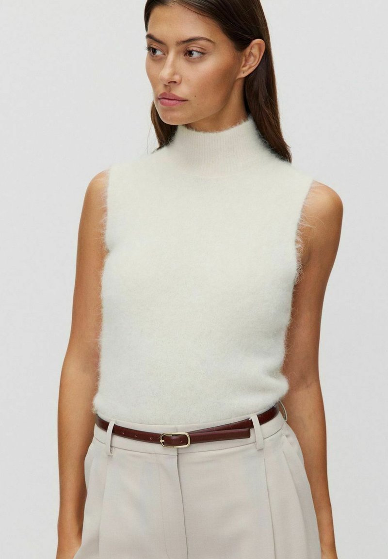 Pull à col roulé sans manches en crème clair avec une texture douce et duveteuse. Associé à un pantalon beige taille haute et une ceinture en cuir marron.