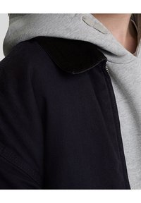 Czarna kurtka z teksturowanej tkaniny i kołnierzem z sztruksu, nałożona na jasnoszary hoodie z regulowanym sznurkiem i miękką fakturą.